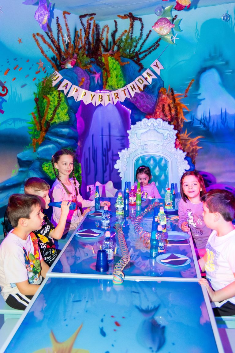 Fröhliche Gruppe von Kindern feiert an einem blau gedeckten Tisch im Unterwasser-Themenraum der Playworld Wien